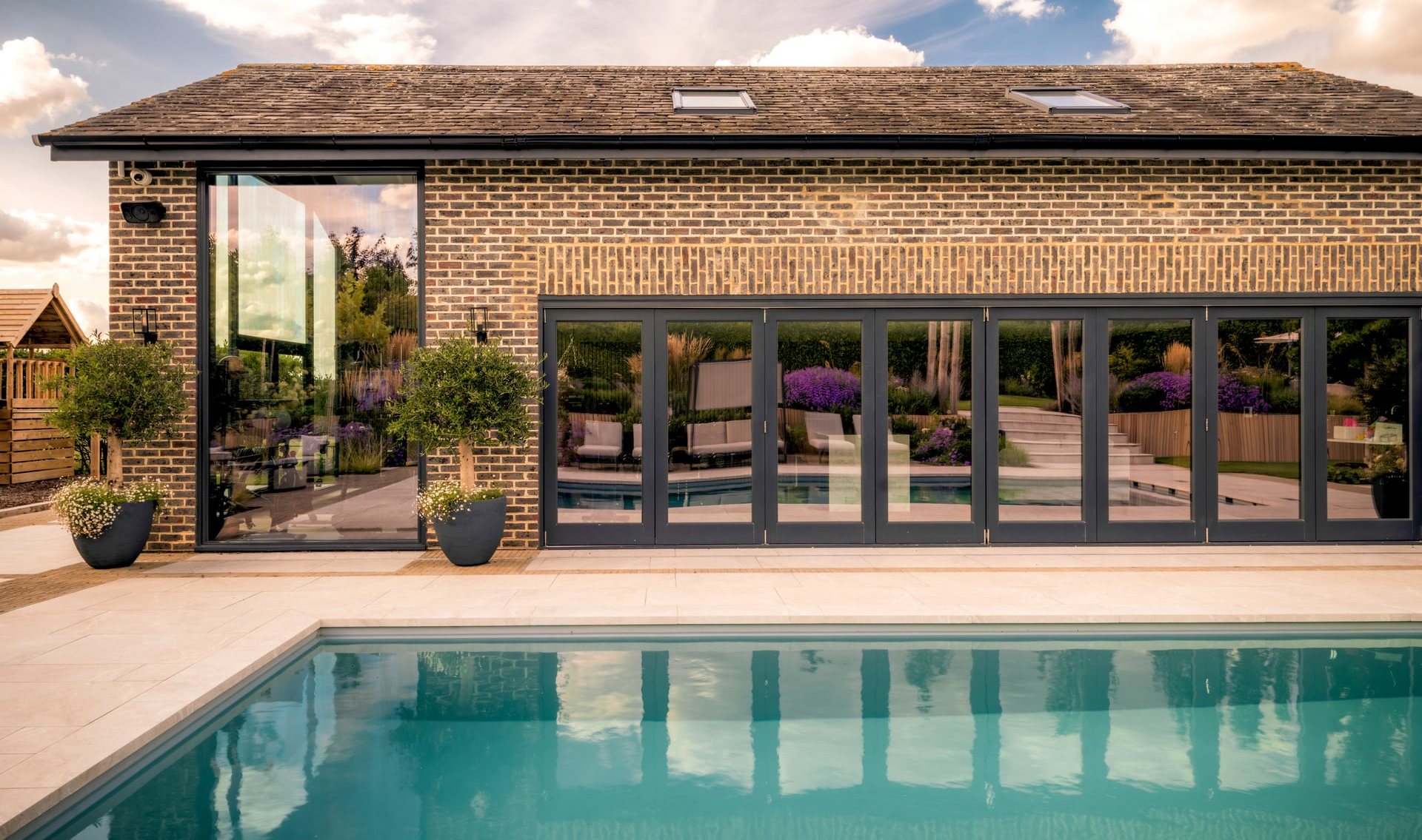 Brick poolhouse and garden room with full-width glazed doors, beside an outdoor swimming pool