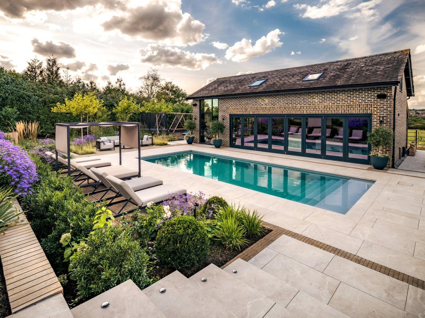 Brick poolhouse and garden room with full-width glazed doors, beside an outdoor swimming pool and planted terrace